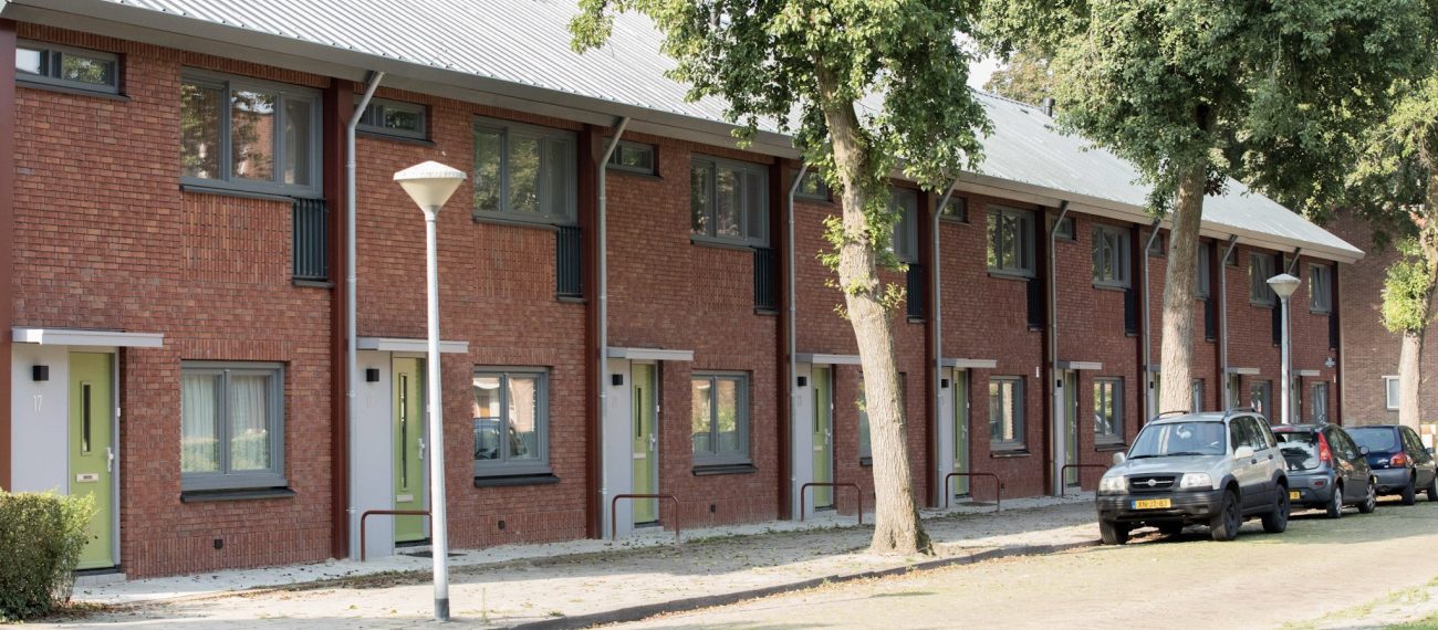 Rij nieuwbouw eengezinswoningen met rode bakstenen gevels en groene voordeuren in een groene woonstraat.