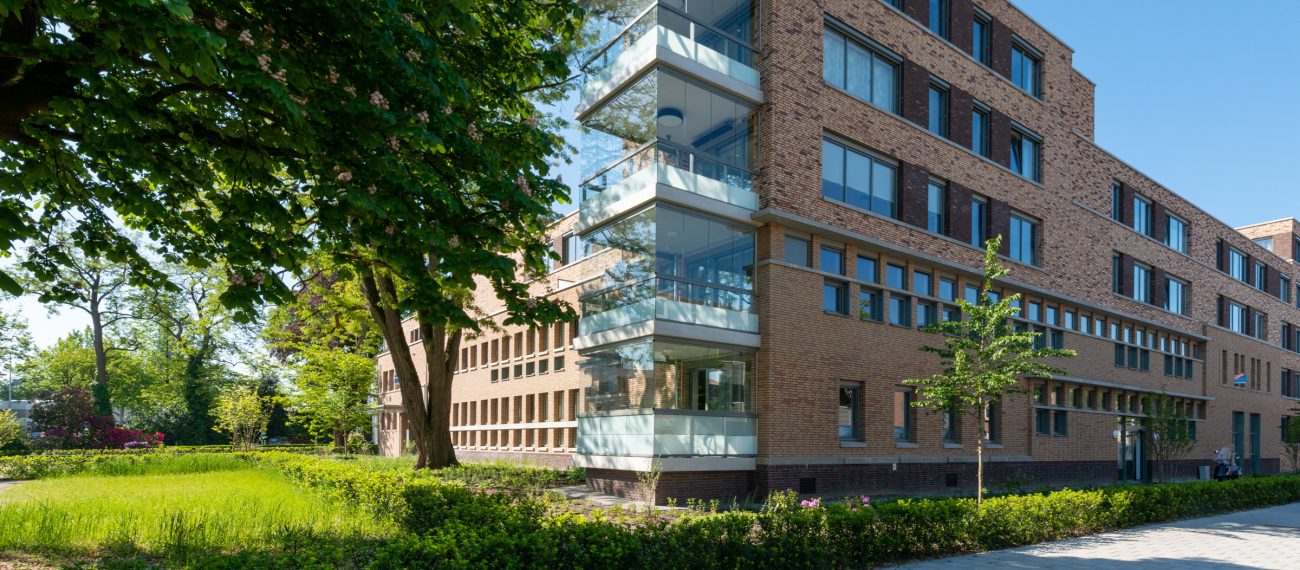 Modern woongebouw met bakstenen gevel en glazen balkons omringd door groen en bomen op een zonnige dag.