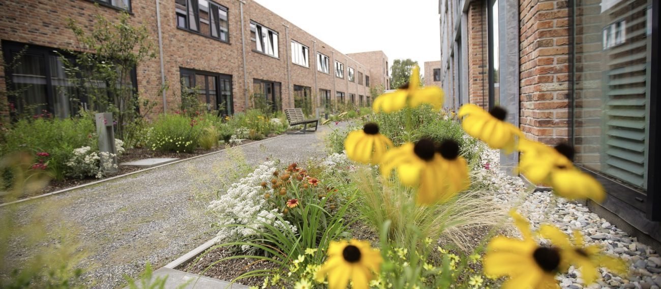 Bloemrijke binnentuin met gele bloemen en groene beplanting tussen lage bakstenen woongebouwen, met wandelpad en zitbank.