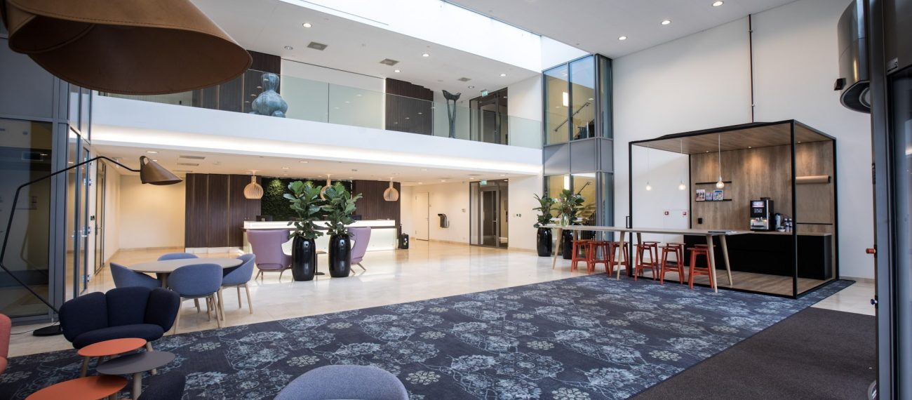 Lichte moderne ontvangsthal met zithoeken, planten, balie en koffiecorner.