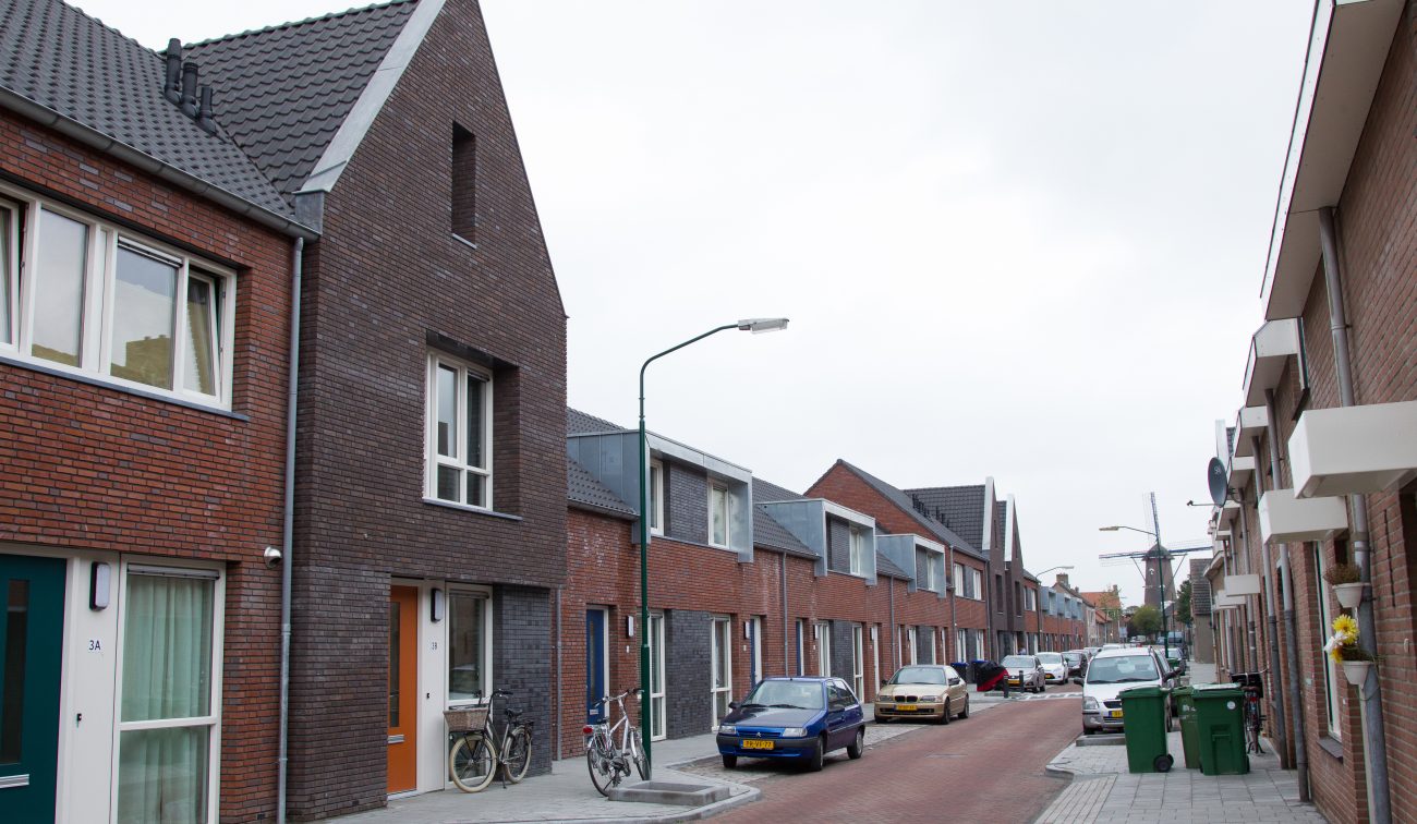 Nieuwbouwwoningen in Zevenbergen met rode en donkergrijze bakstenen gevels, oranje voordeuren en moderne zinken dakkapellen, gelegen aan een smalle woonstraat met geparkeerde auto's en fietsen.