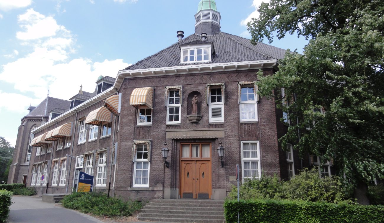 Verbouwing Huize Padua Boekel – monumentale hoofdentree met houten deuren, bakstenen gevel, witte kozijnen en kleine toren, gezien vanaf de oprijlaan met groen.