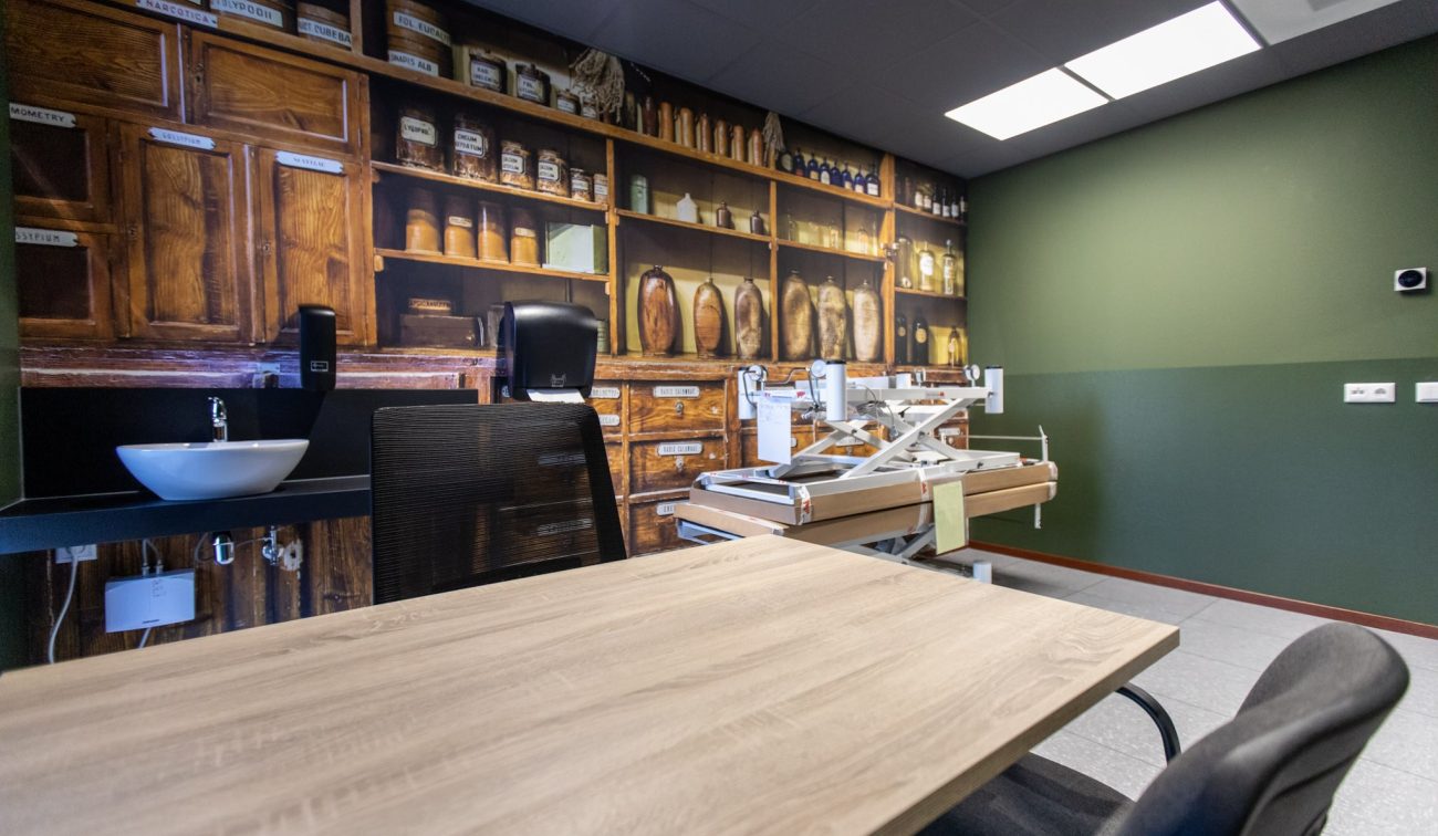 Interieur behandelruimte – groene wand met apothekerskast-muurprint, wastafel en verstelbare behandeltafel naast bureau