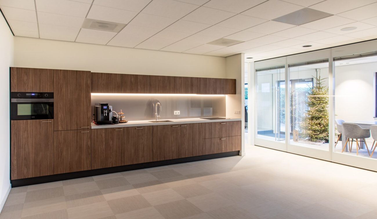 Moderne pantry met houten kasten, inbouwoven, koffieautomaat en spoelbak met onderkastverlichting; rechts glazen schuifdeuren naar vergaderruimte met kerstboom.