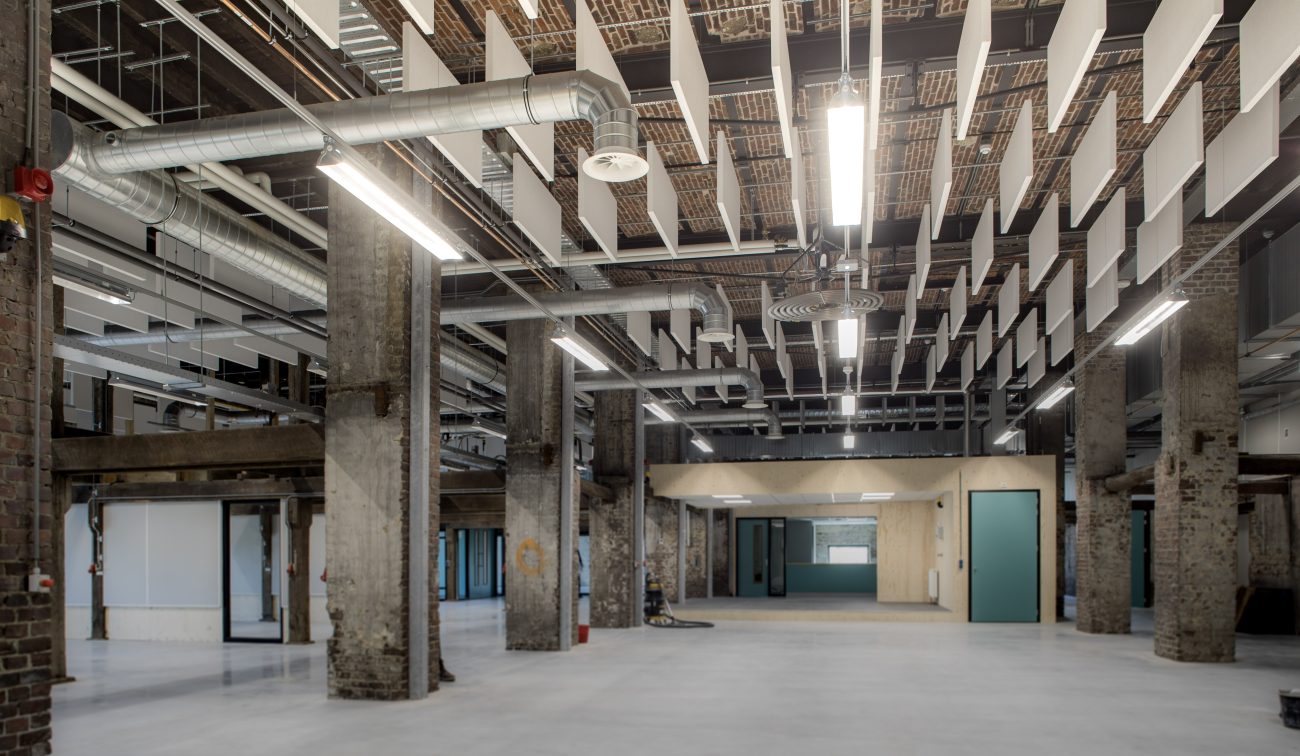Industrieel interieur van de getransformeerde Greswarenfabriek: open hal met ruwe betonnen kolommen, zichtbaar leidingwerk en hangende akoestische panelen, gladde betonvloer en mintgroene deuren.