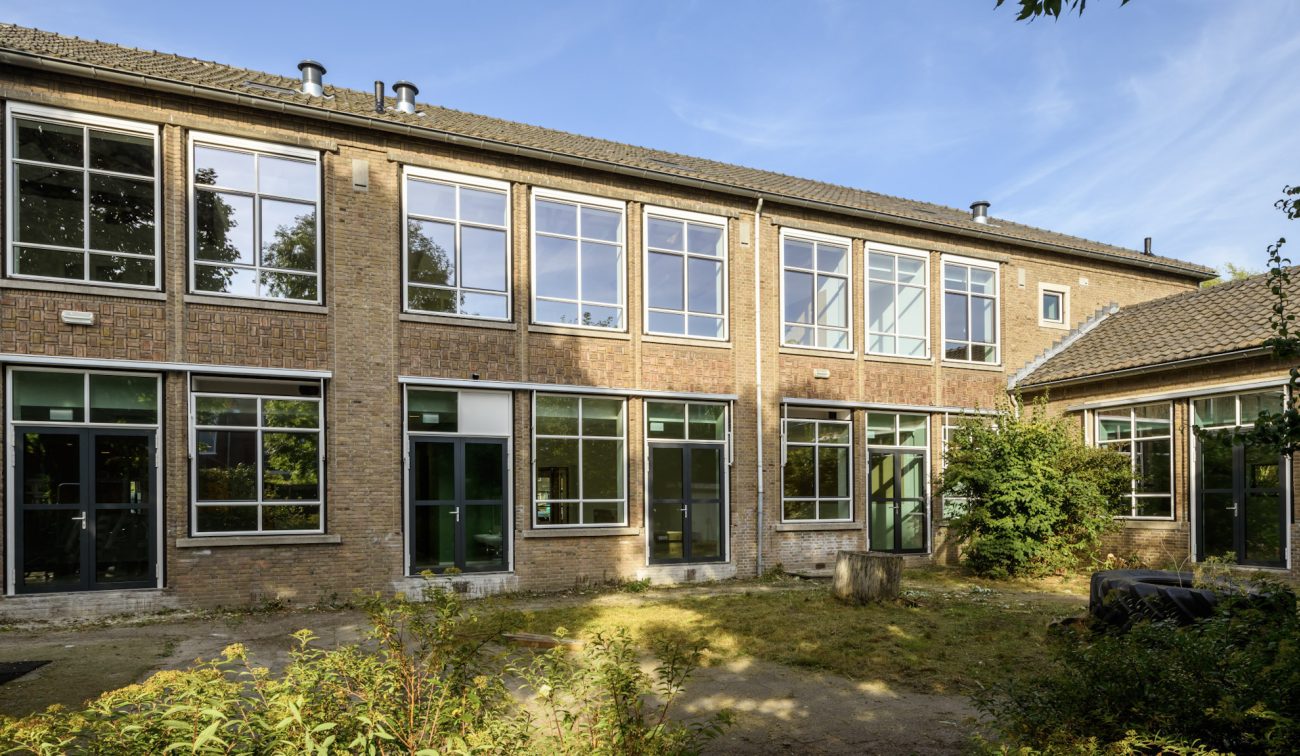 Gevel OBS De Fakkel: twee lagen baksteen met hoge stalen ramen, groene deuren en binnentuin