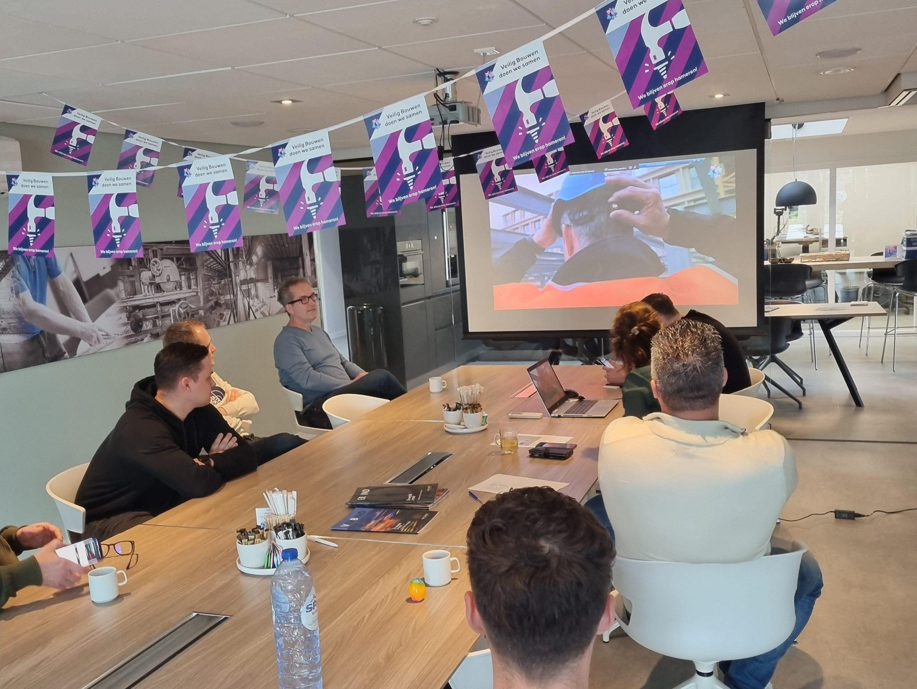 Veiligheidsbijeenkomst met presentatie rond vergadertafel en vlaggenlijn over veilig bouwen