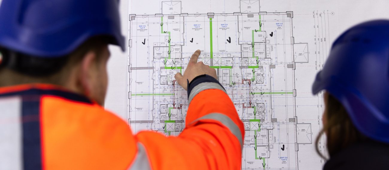 Twee medewerkers met helmen in veiligheidshesjes bekijken en bespreken een technische bouwtekening, terwijl één persoon naar een onderdeel op het plan wijst.