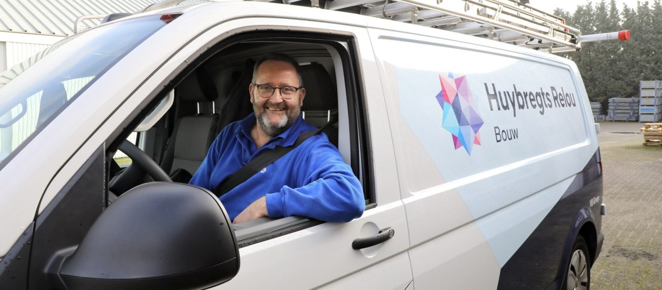 Man zit vrolijk kijkend achter het stuur van een bedrijfsbus met het raam open en zijn arm uit het raam