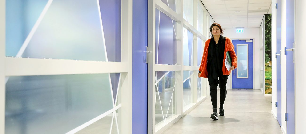 Medewerker loopt door modern kantoor met glazen wanden en blauwe deuren, documenten onder de arm