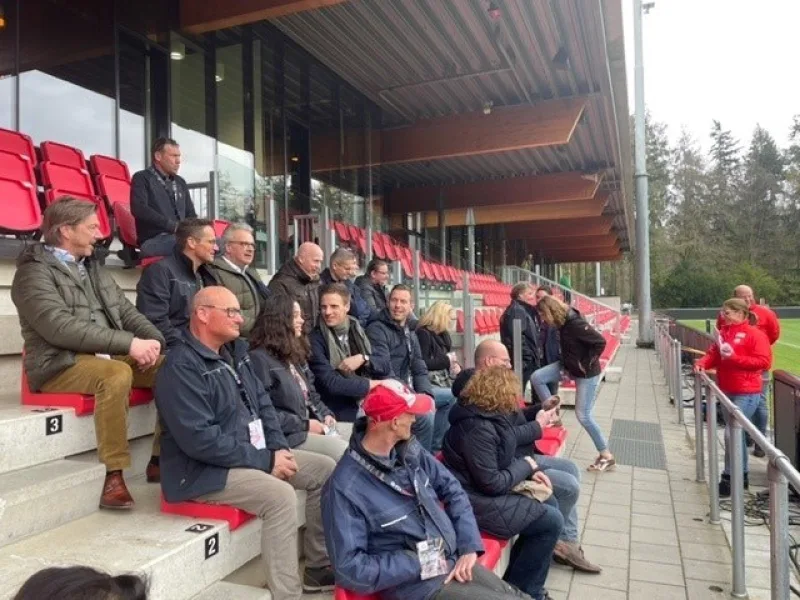 Groep bezoekers op rode tribune tijdens bijeenkomst bij voetbalveld