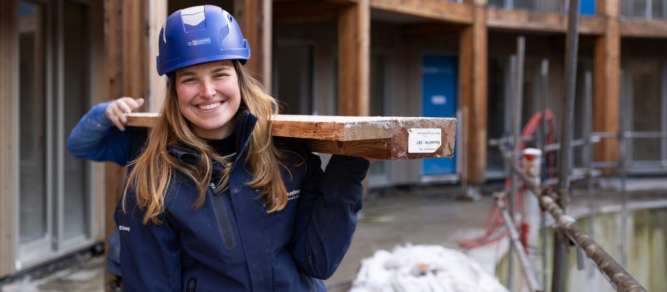 Vrouw met blauwe bouwhelm glimlacht terwijl ze een houten balk op haar schouder draagt op een bouwplaats.
