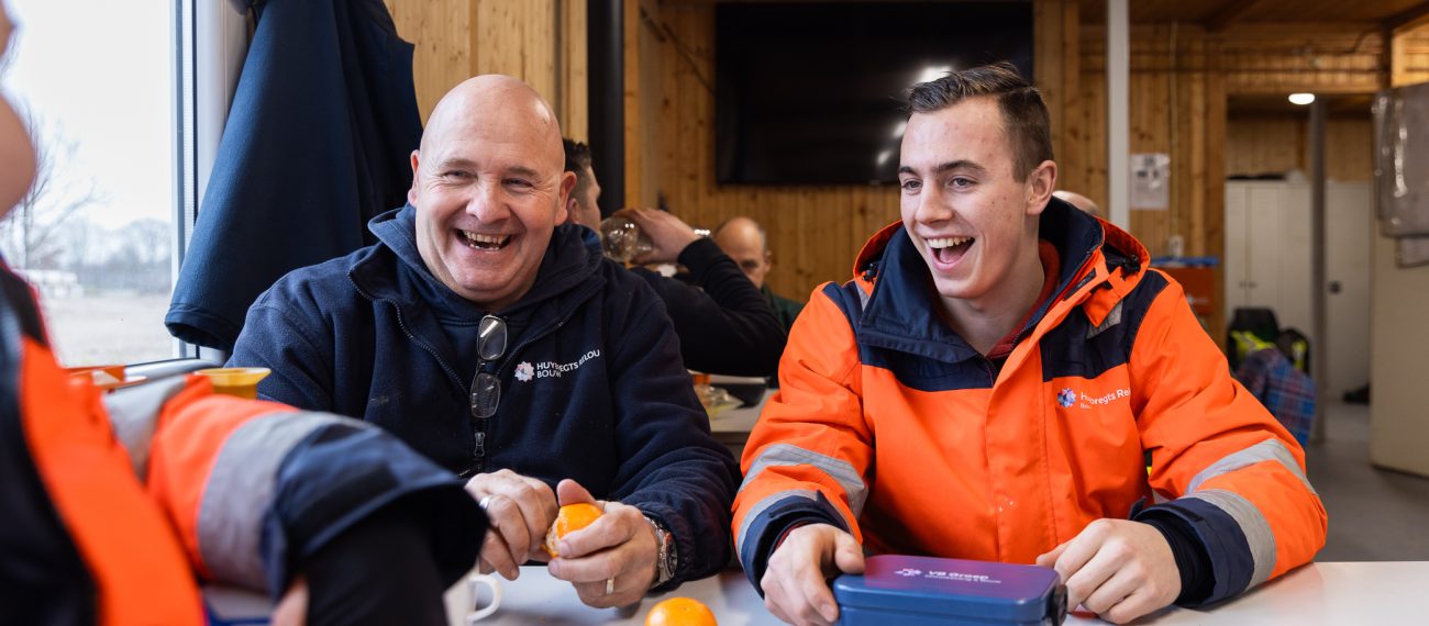 Twee bouwmedewerkers lachen samen tijdens de pauze aan een tafel.