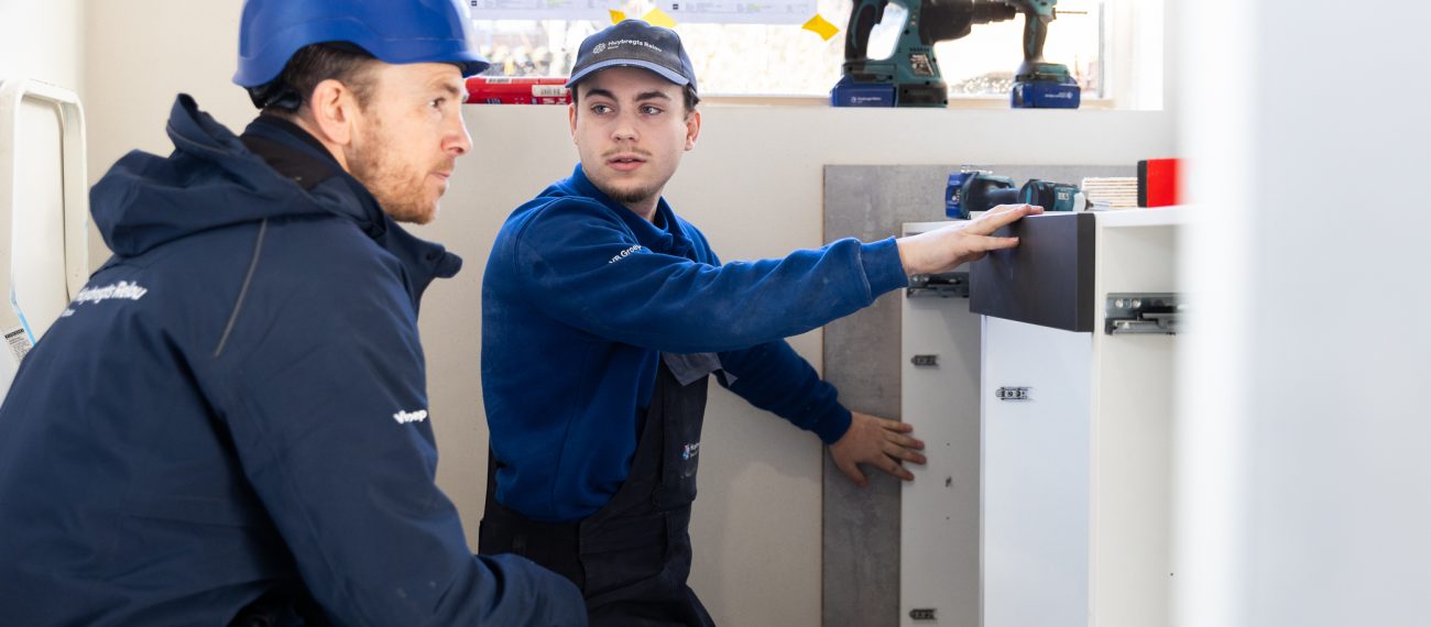 Twee medewerkers met beschermende kleding en helmen installeren onderdelen in een interieur; één persoon geeft uitleg terwijl de ander aan een kast werkt met gereedschap op de achtergrond