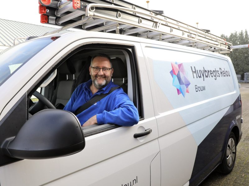 Man zit vrolijk kijkend achter het stuur van een bedrijfsbus met het raam open en zijn arm uit het raam