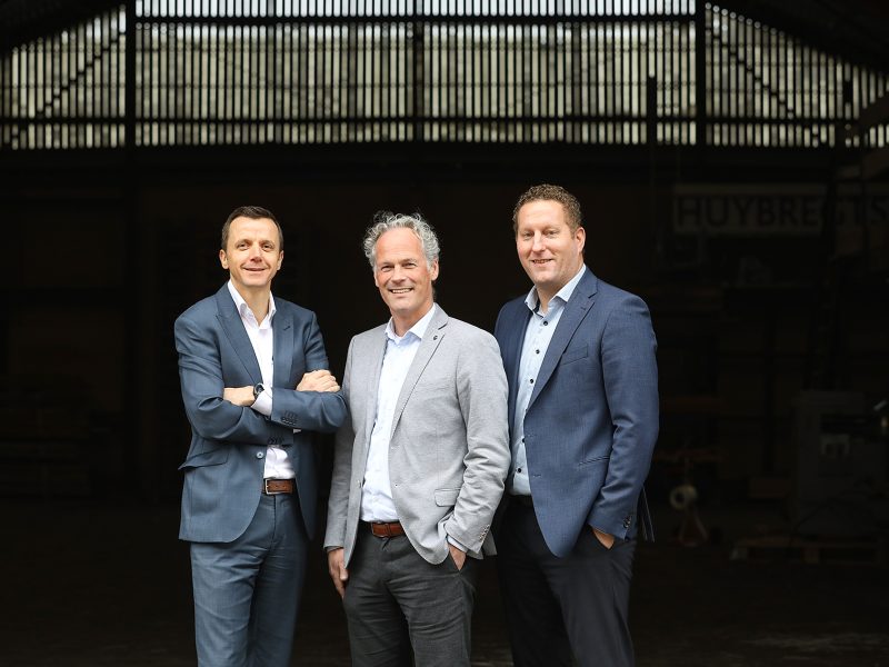 Drie mannen in pak staan naast elkaar en poseren lachend voor een donkere industriële achtergrond.