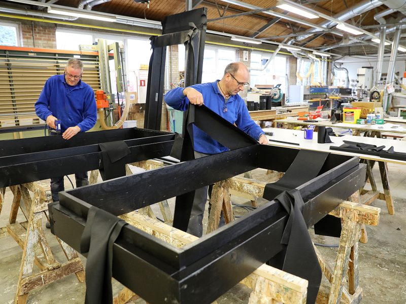 Twee mannen in een werkplaats bevestigen zwarte materialen aan houten frames op werkbanken.