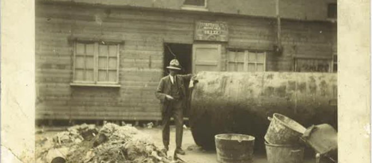 Man naast grote metalen tank en puin voor houten gebouw in industriële setting, historische foto
