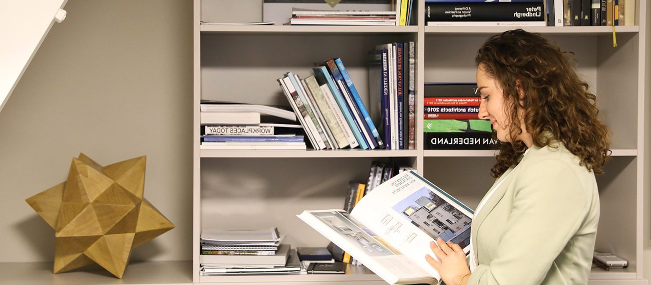 Vrouw bekijkt vastgoedboek in kantoor, boekenkast met vakliteratuur en houten designobject op achtergrond.