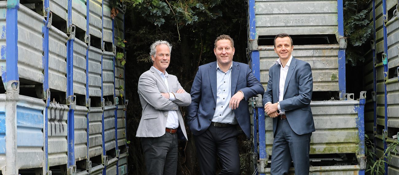 Drie mannen in pak poseren buiten tussen gestapelde metalen bakken en kijken lachend naar de camera.