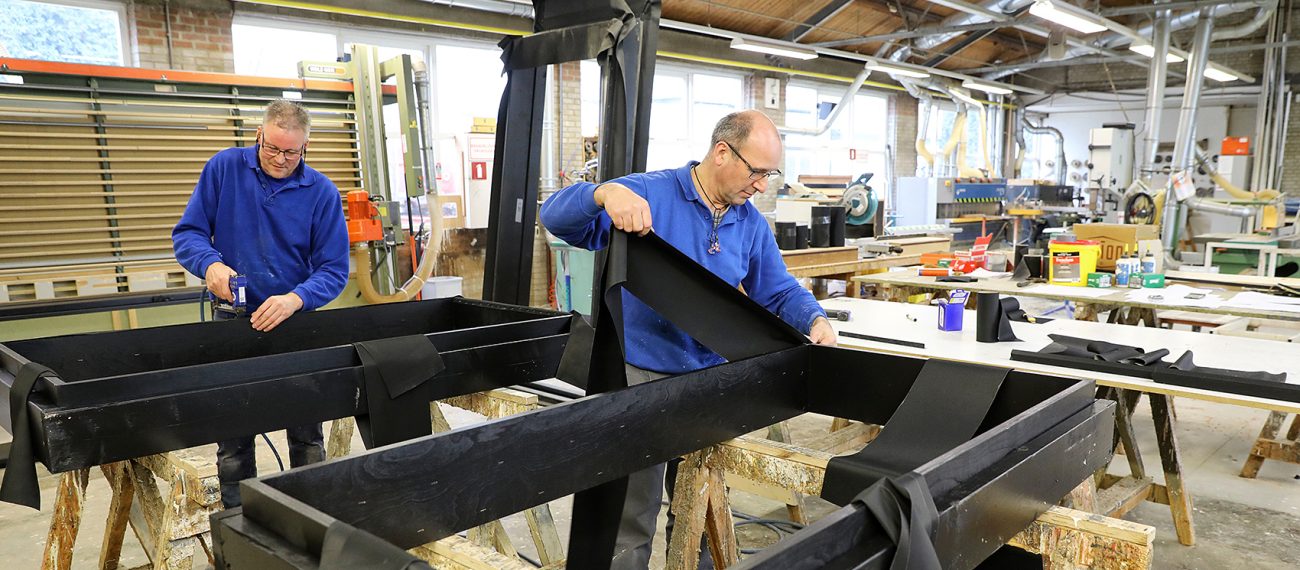 Twee medewerkers werken in een houtwerkplaats aan grote zwarte houten constructies; één bevestigt onderdelen met een gereedschap terwijl de ander zwarte banen materiaal plaatst.