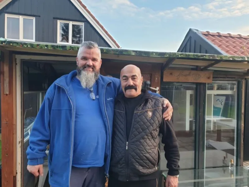 Twee mannen met gezichtsbeharing staan samen voor een woning met houten gevel en glazen pui