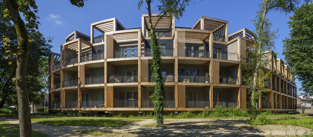Rond woongebouw met meerdere verdiepingen en houten gevels, omringd door bomen en groen.