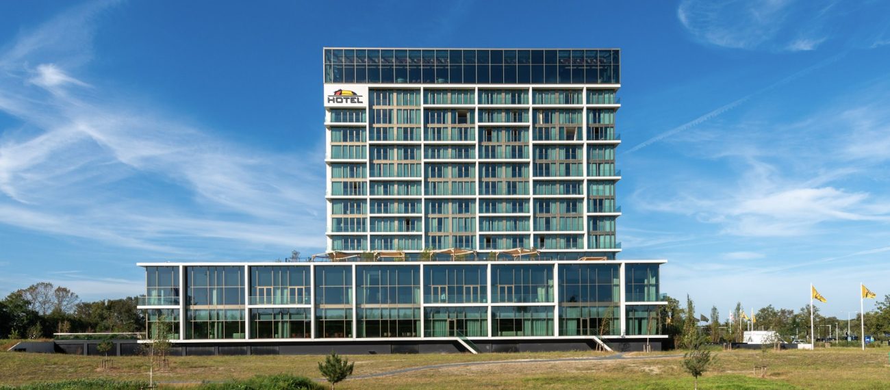 Modern hoog hotelgebouw met glazen gevel, groene accenten en meerdere verdiepingen, gefotografeerd frontaal onder een heldere blauwe lucht met grasveld op de voorgrond.