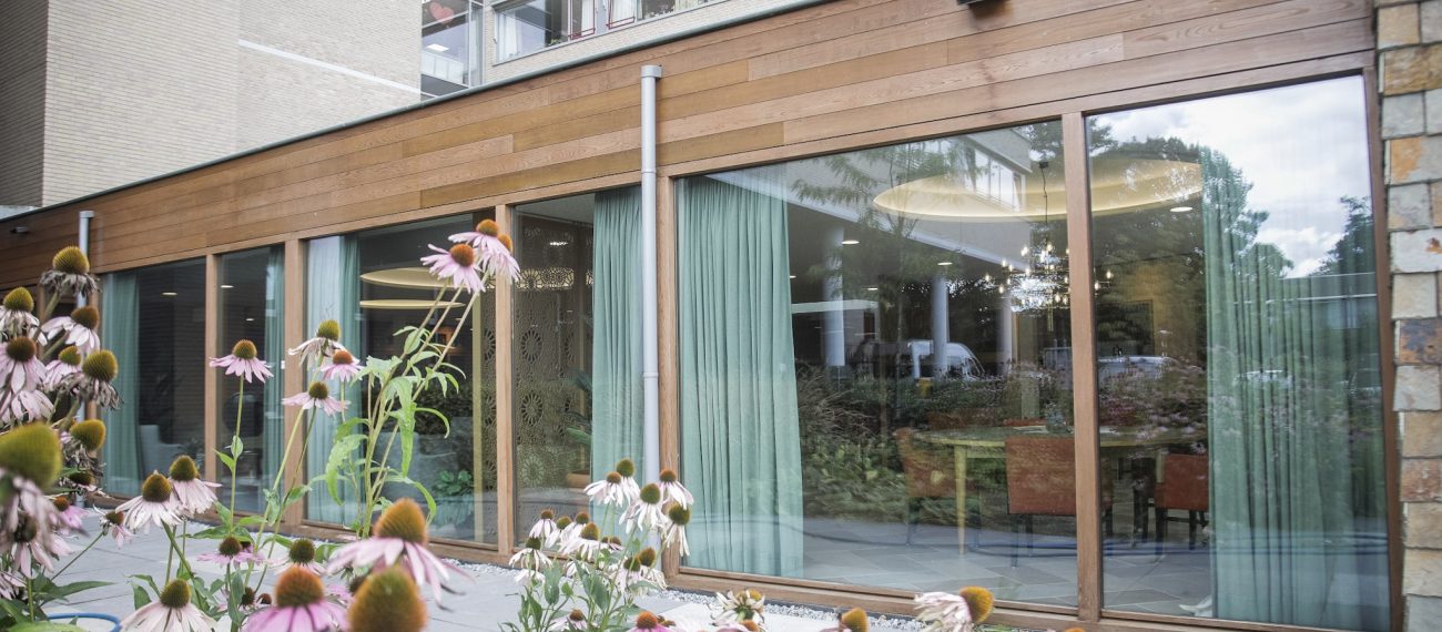 Gevel met houten afwerking en grote ramen met bloemen op de voorgrond.