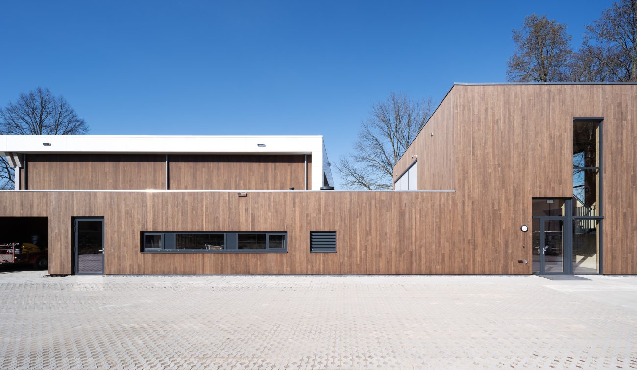 : Voorgevel van de nieuwbouw Brandweerpost Sittard-Oost met lange, horizontale houtbeklede volumes, een open garagebox links, smalle raamstrook en hoge glazen entree rechts onder een strakblauwe lucht.