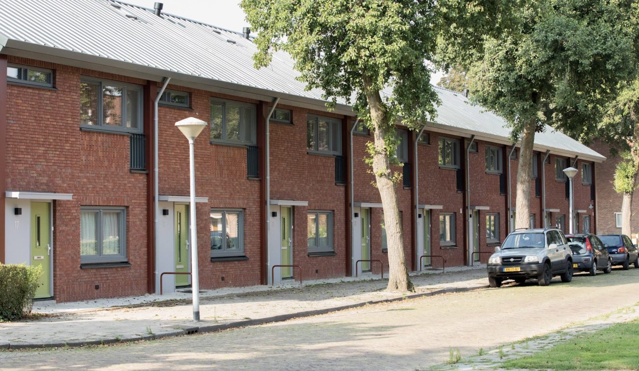 Renovatie (groot onderhoud) NOM van 195 rijwoningen in ’t Ven, Eindhoven – straatbeeld vanaf beneden met rode baksteen, groene voordeuren, nieuw metalen dak en bomen langs de stoep.