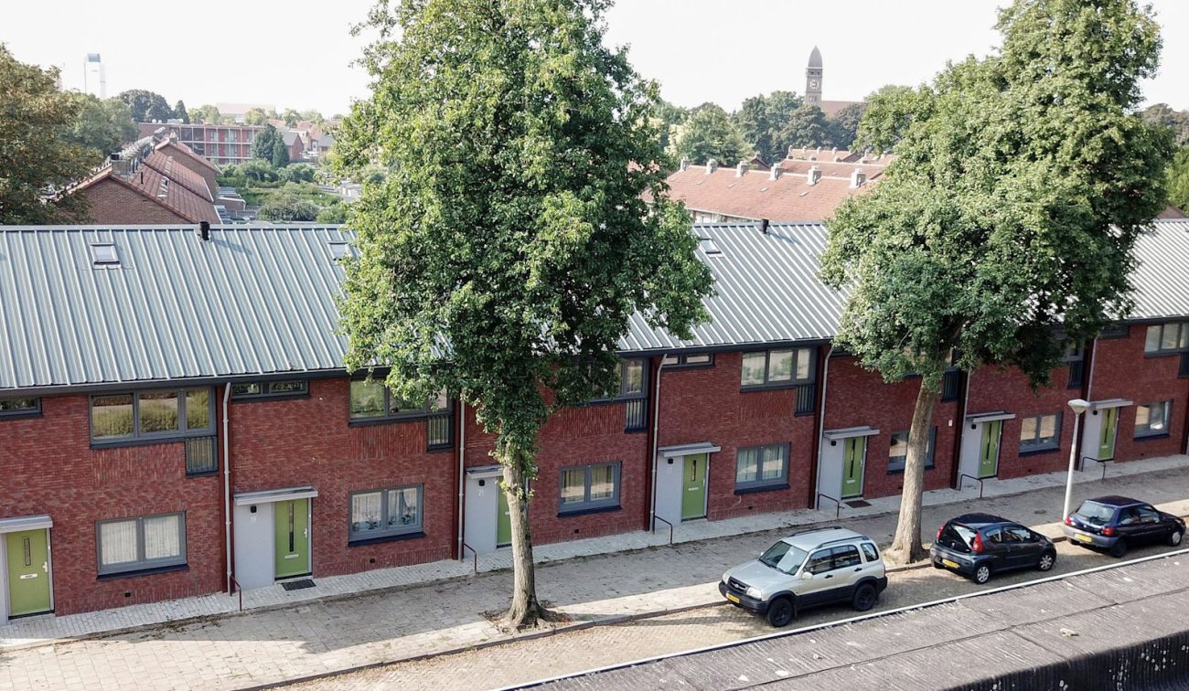 Renovatie (groot onderhoud) NOM van 195 rijwoningen in ’t Ven, Eindhoven – overzicht vanaf hoogte van een rij woningen met rode baksteen, groene voordeuren en nieuw metalen dak aan een boomrijke straat met geparkeerde auto’s.