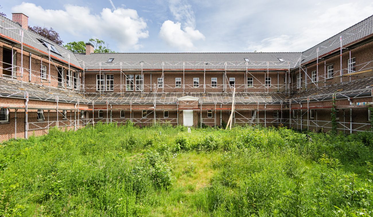 Binnenplaats van bakstenen gebouw met steigers rondom en begroeid binnenterrein tijdens renovatie