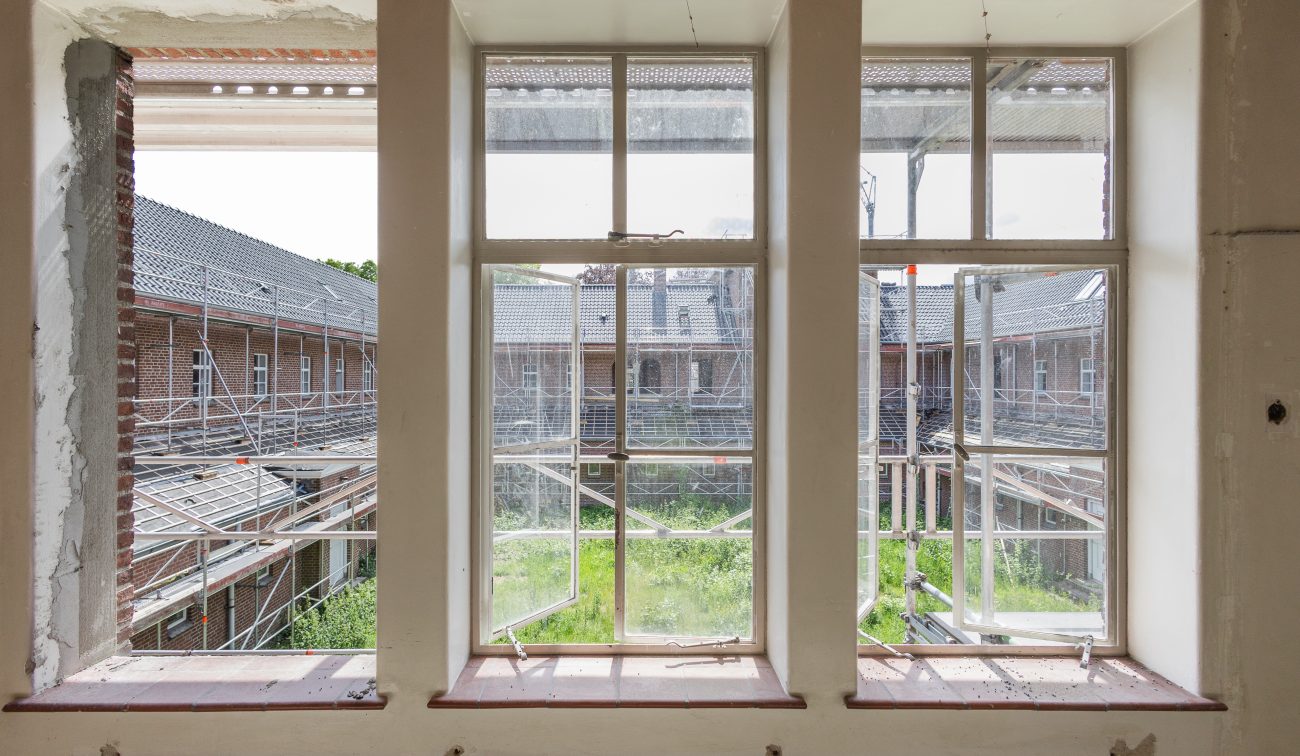Uitzicht door ramen op steigers en begroeide binnentuin tijdens renovatie van bakstenen gebouw