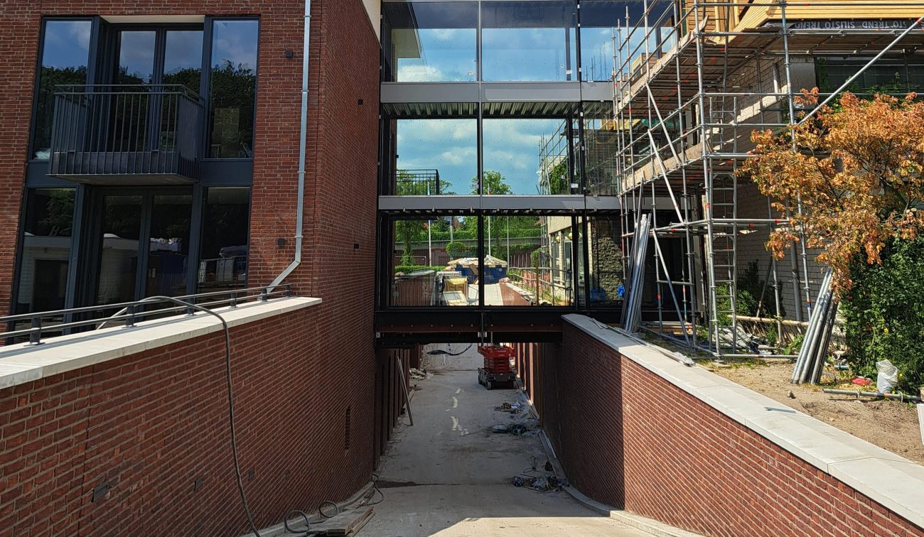 Uitbreiding en renovatie Van der Valk Hotel Wassenaar – glazen loopbrug die twee hotelvleugels verbindt boven de inrit van de parkeergarage, met bakstenen gevels, zinken daken en steigers in de bouwfase.