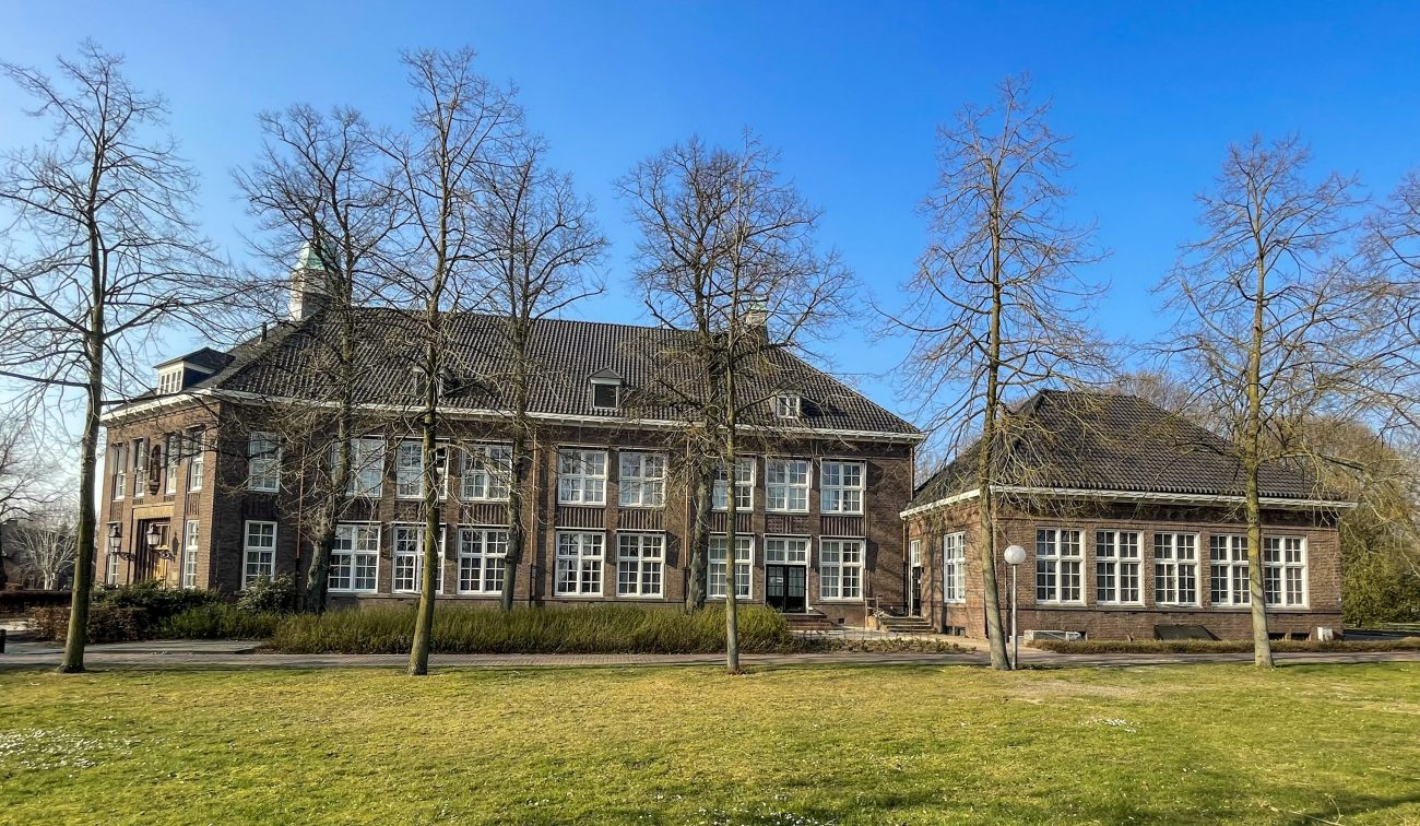 Verbouwing Huize Padua in Boekel – monumentaal gebouw met klassieke bakstenen gevels en hoge ramen, gezien vanaf het park met bomen en grasveld onder een heldere blauwe lucht.