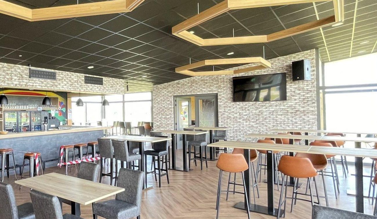 Interieur kantine clubgebouw DVO’20 met bar, hoge en lage tafels, oranje barkrukken, hexagonale houten plafondframes en visgraatvloer; grote tv aan de muur.