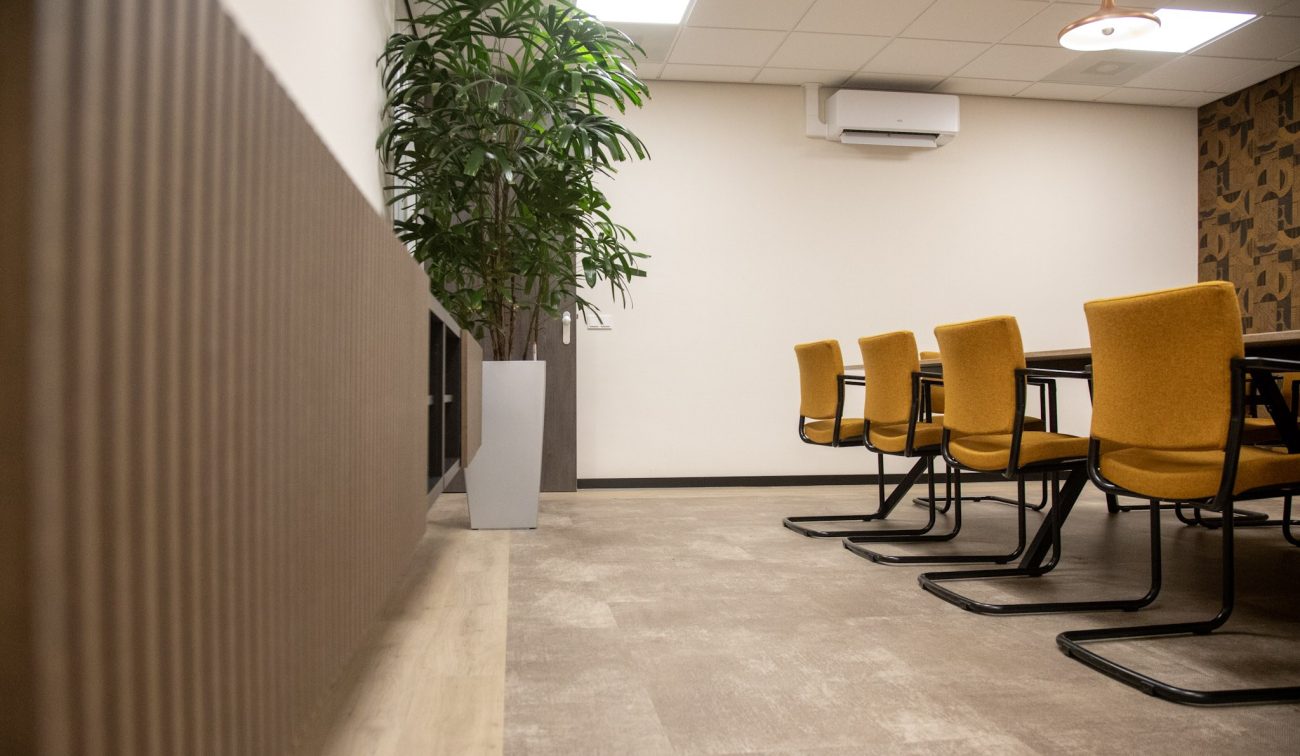 Vergaderruimte met mosterdgele sledestoelen aan een tafel, een hoge kamerplant in een witte pot, lattenwand links, airco-unit hoog aan de muur en neutrale pvc-vloer onder systeemplafond.