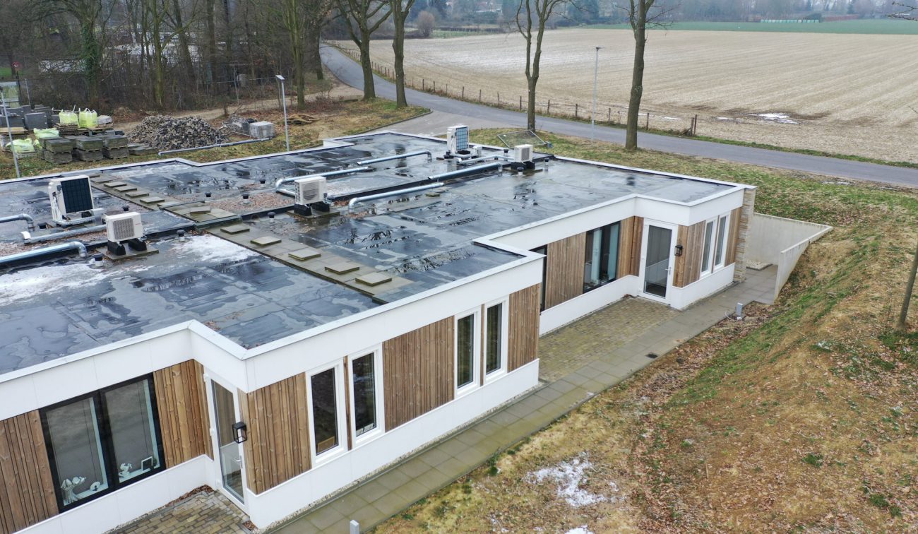 Hoekaanzicht nieuwbouw zorgwoning Amstenrade met witte en houten gevel, plat dak vol installaties, betegeld pad langs de gevel en landelijke omgeving met weg en velden.
