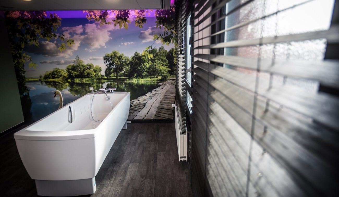 Zorgbadkamer met hoog ligbad, houtlook vloer en natuurwand met meerzicht voor rust en beleving