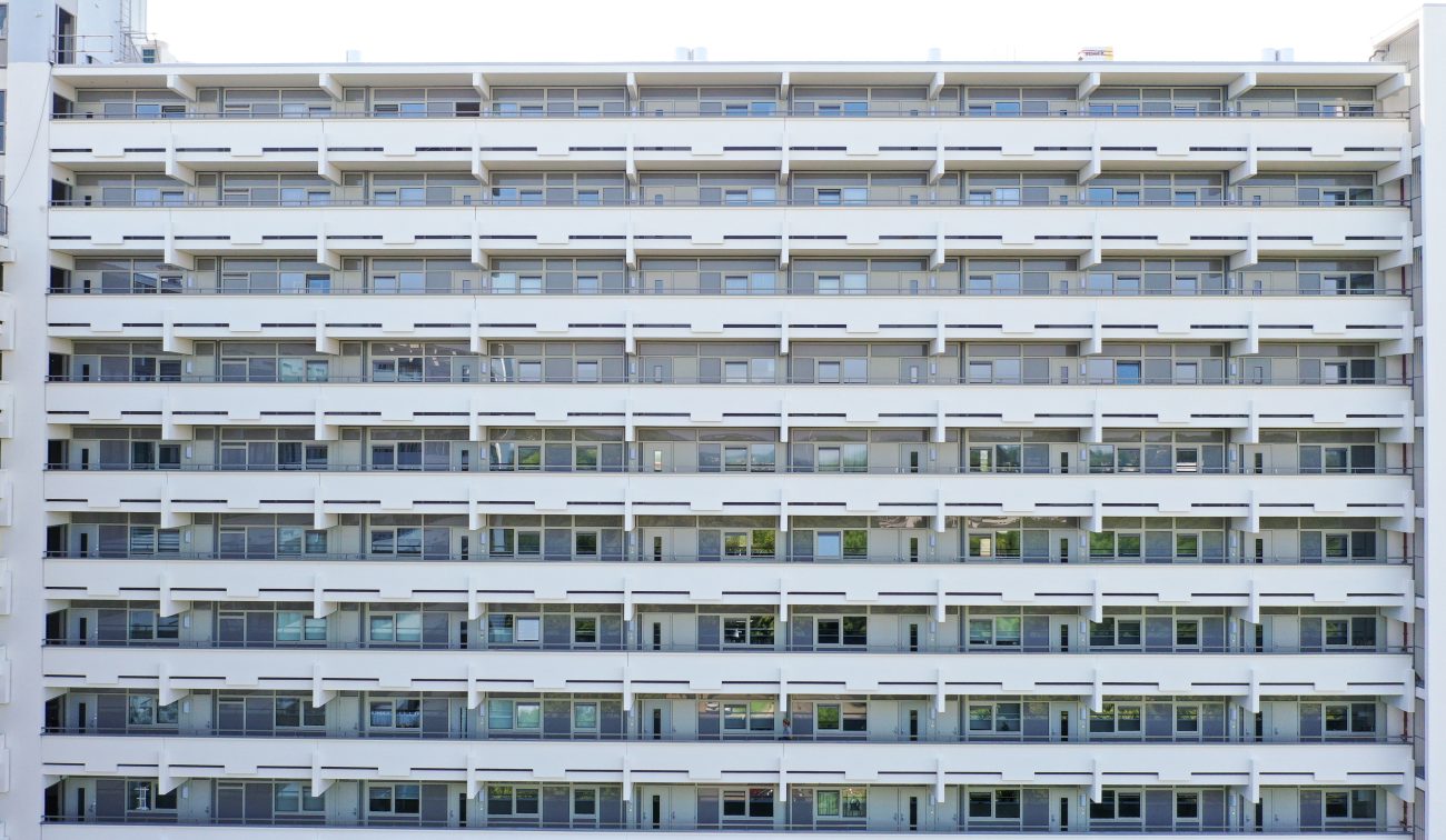 Strakke witte galerijflat in frontaal aanzicht met repetitieve horizontale lijnen van balkons en ramen over meerdere verdiepingen.