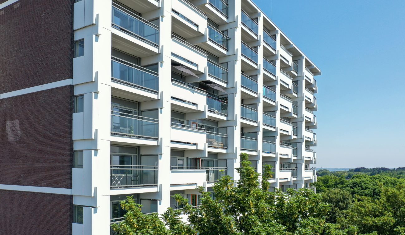Close-up van de gerenoveerde galerijflat met witte gevelelementen en glazen balkonhekken; bakstenen kopgevel links, bomen en blauwe lucht op de achtergrond.