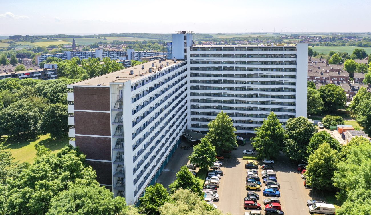 Luchtfoto van L-vormige galerijflat Veliahof in Maastricht met witte gerenoveerde gevels en lange balkonstroken, omringd door bomen en parkeerplaatsen.