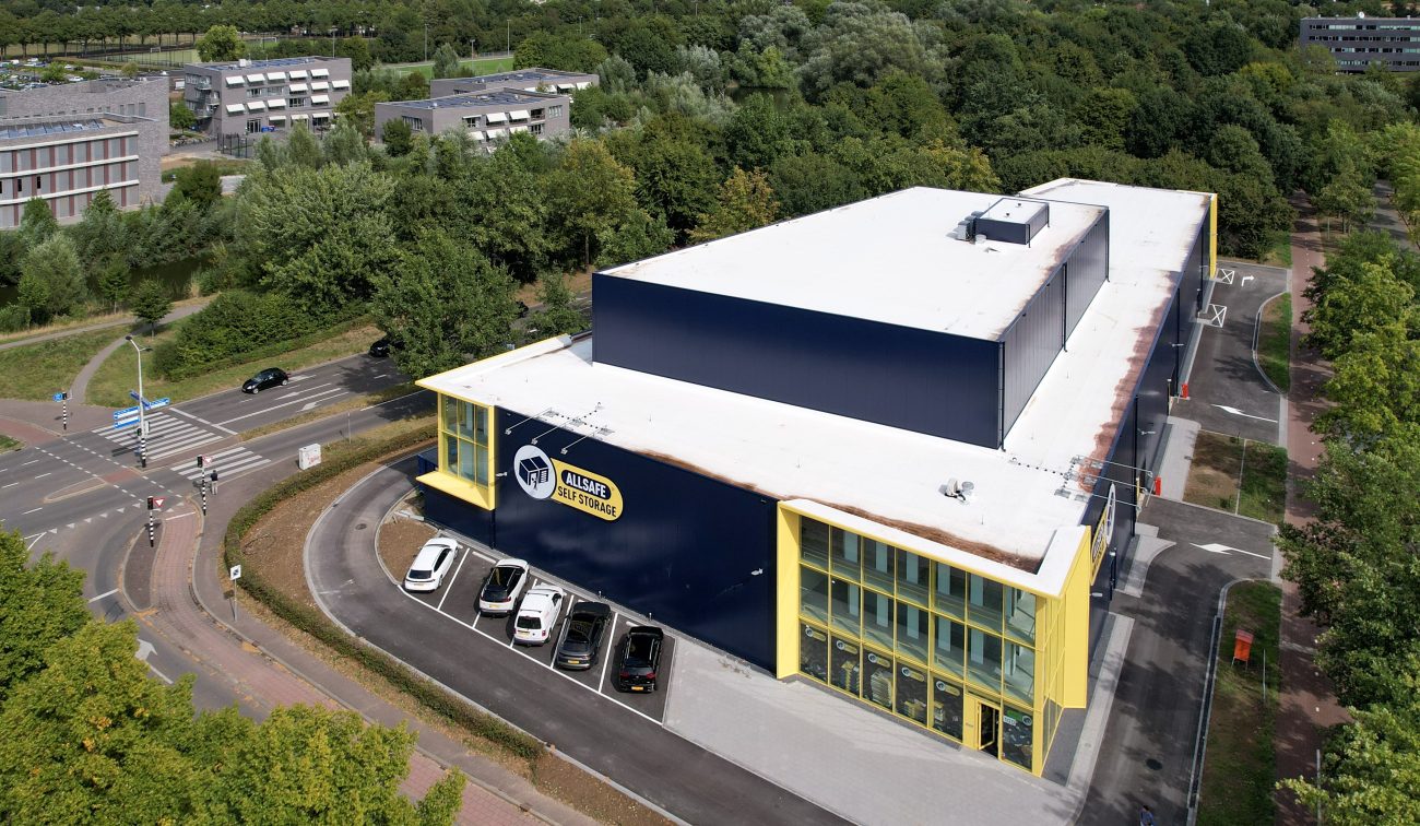 Luchtfoto van de nieuwbouw van Allsafe Self Storage in Maastricht: donkerblauw gebouw met witte dakplaat en felgele kaders rond de glazen hoekgevel, kleine parkeerstrook met auto’s langs de gevel, gelegen aan een kruising en omringd door bomen.