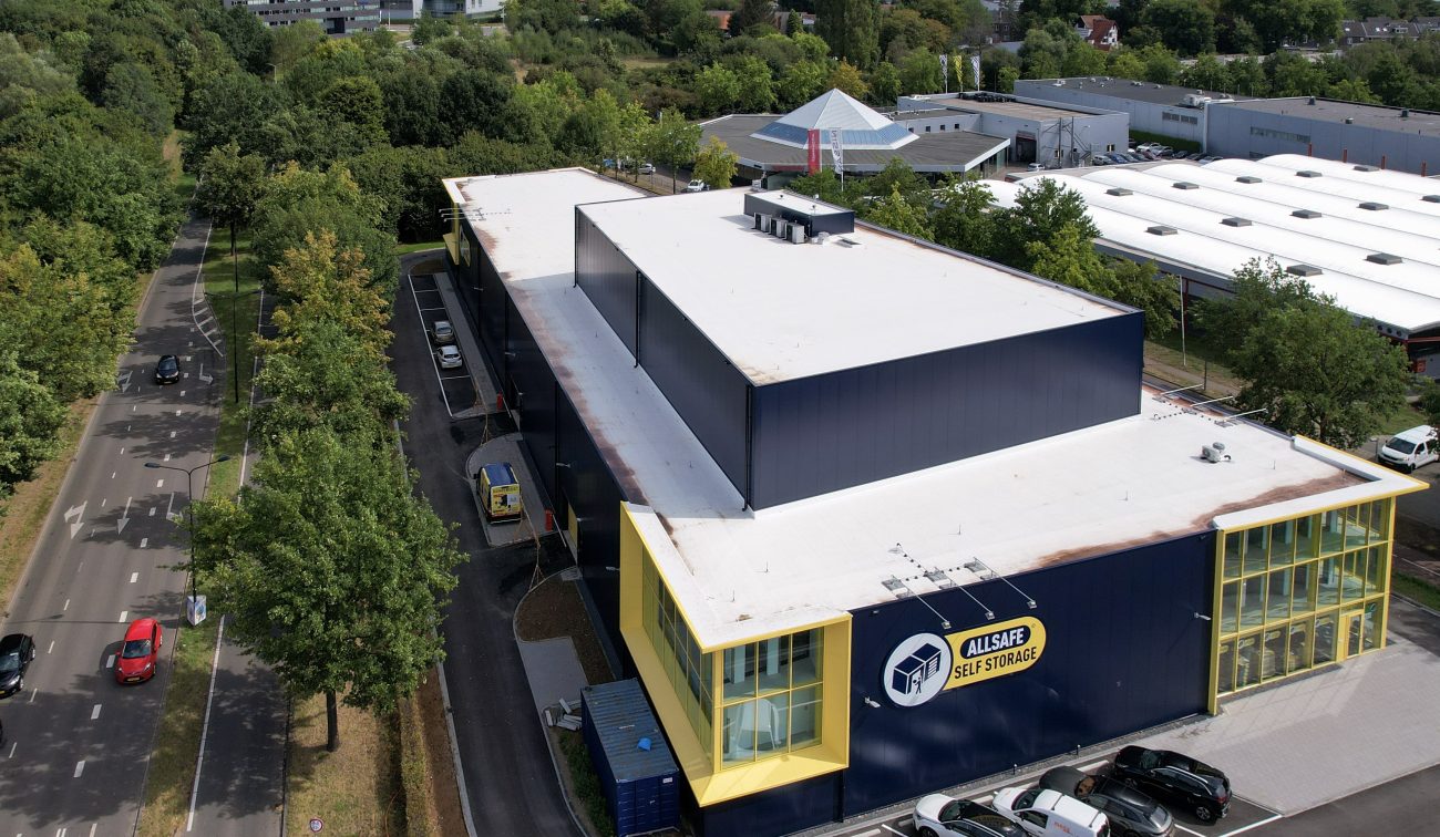 Luchtfoto van de nieuwbouw Allsafe Self Storage in Maastricht: rechthoekig donkerblauw opslaggebouw met wit dak en geel omlijnde glazen hoeken, logo op de gevel, smalle inrit met geparkeerde auto’s en bestelbus, bomen langs de weg en lage bedrijfsgebouwen in de omgeving.