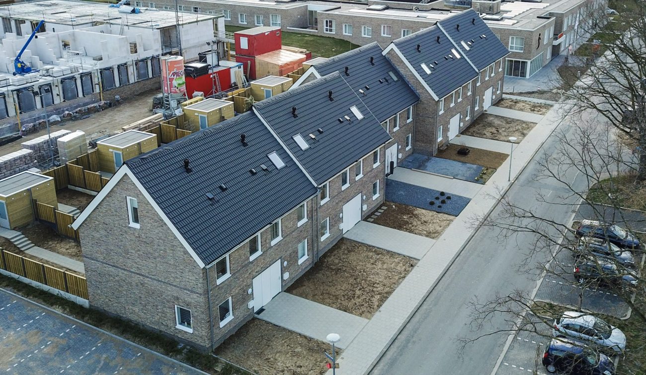 Nieuwbouw rij eengezinswoningen in Hulsberg met bakstenen gevels en donkere pannendaken; nieuwe stoepen en opritten, houten tuinen/bergingen en bouwplaats links.