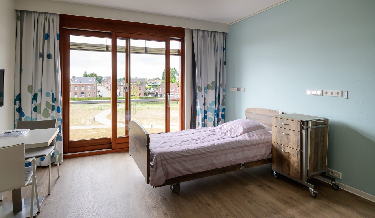 Verpleeghuiskamer met houten vloer, bed op wielen en gordijnen met blauw patroon.
