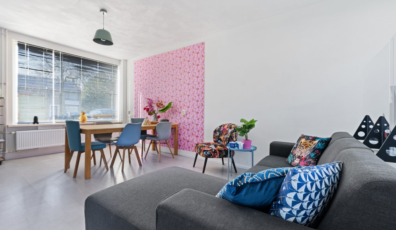 Moderne woonkamer met grote ramen, houten eettafel, mix stoelen, grijze bankje en roze accentbehang.