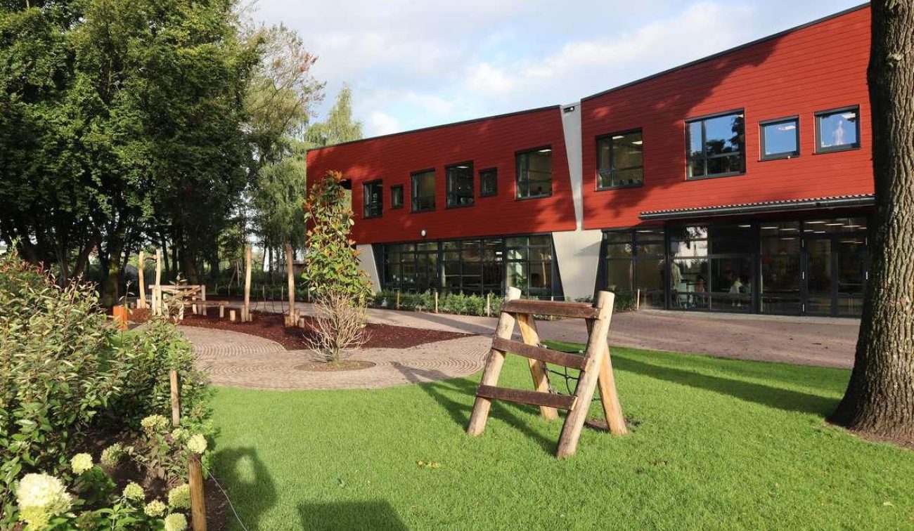 Groen schoolplein met gazon en natuurlijke speeltoestellen van hout; op de achtergrond de rood beklede gevel van basisschool Den Doelhof met grote ramen en glazen entree.