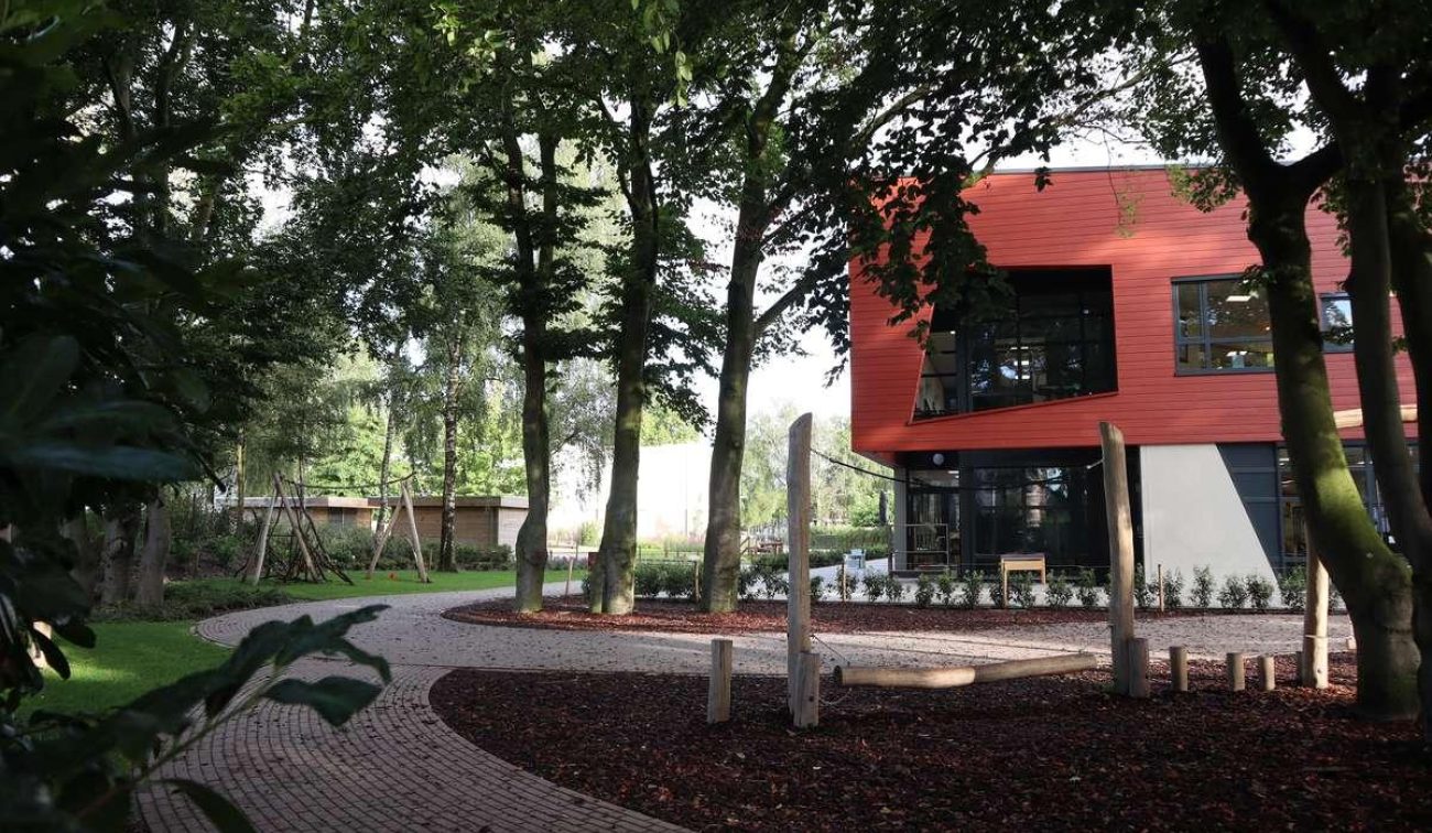 Groen schoolplein met slingerend pad tussen hoge bomen; rechts de rood beklede gevel van basisschool Den Doelhof met grote ramen en een natuurlijk speelgedeelte met houten palen.